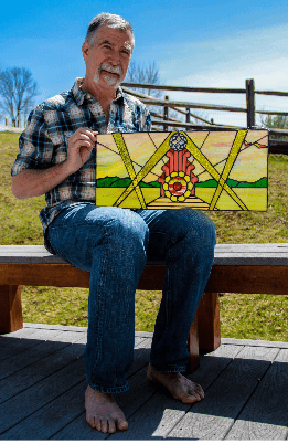 Barefoot Andy, sitting on bench holding up completed stained glass window "Showtime"
