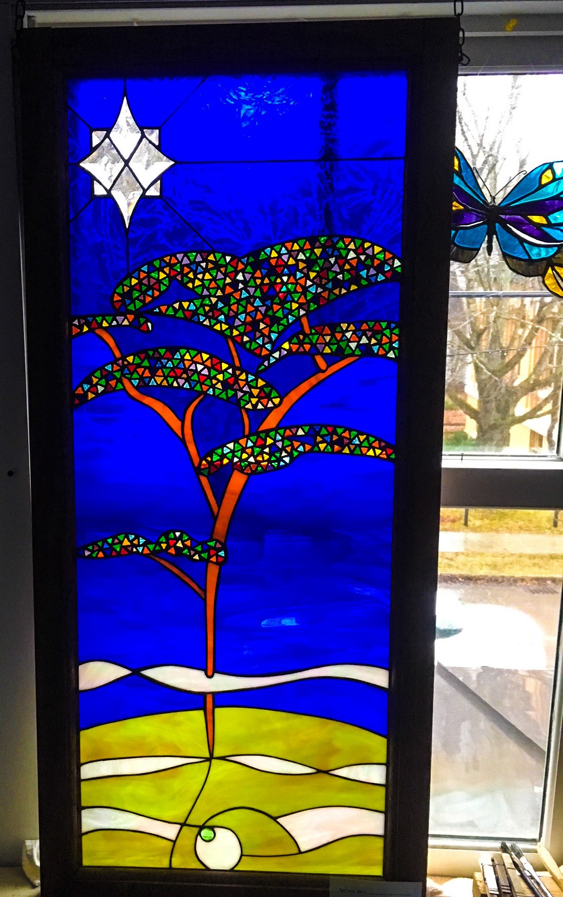 Stained glass image of a tree reaching to the heavens from Earth below. Blue star-lit sky, green earth below, multi-colored tree branches.