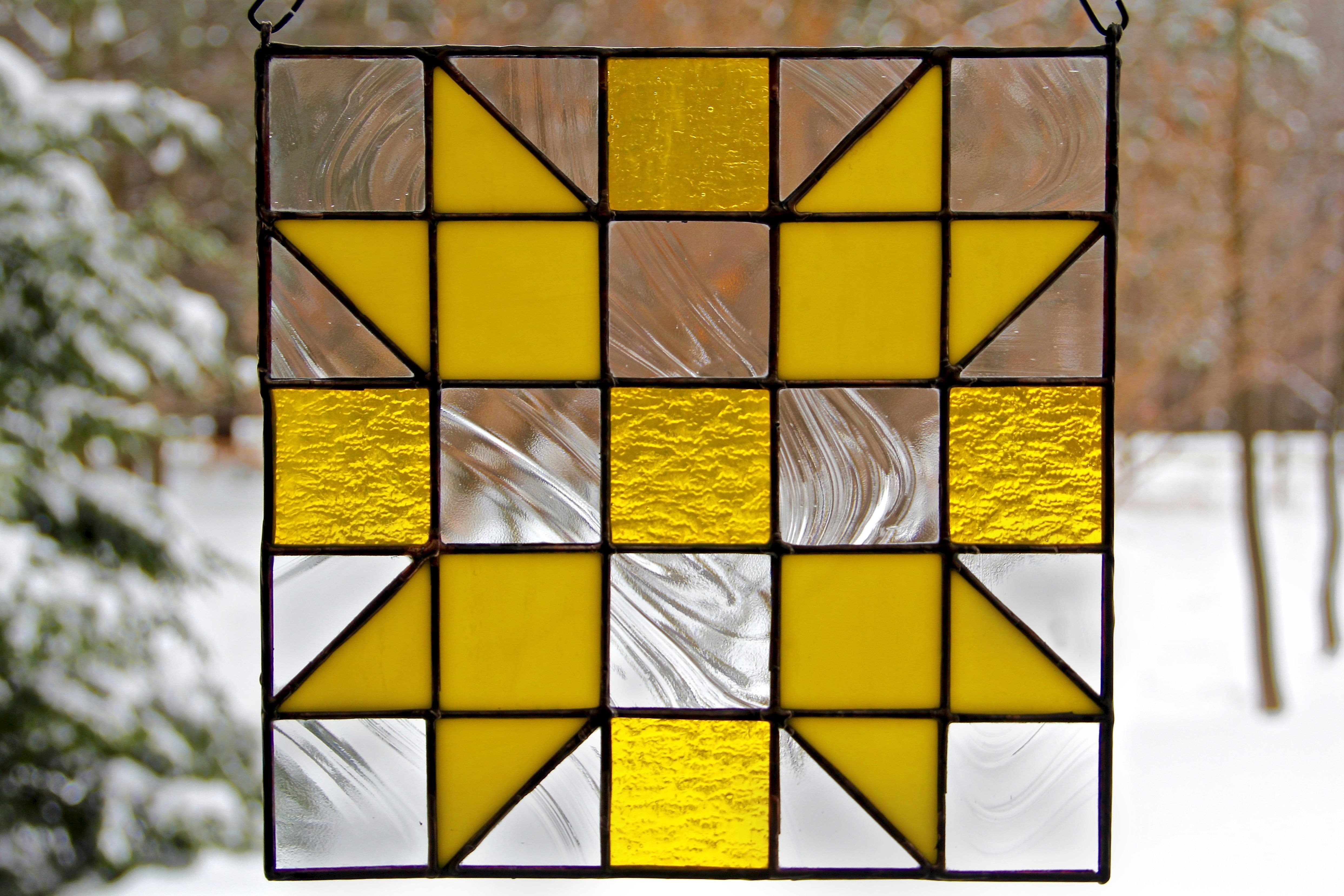 Stained glass rendition of "Sisters," a single block quilt pattern, in yellow, against snowy wooded scenery.